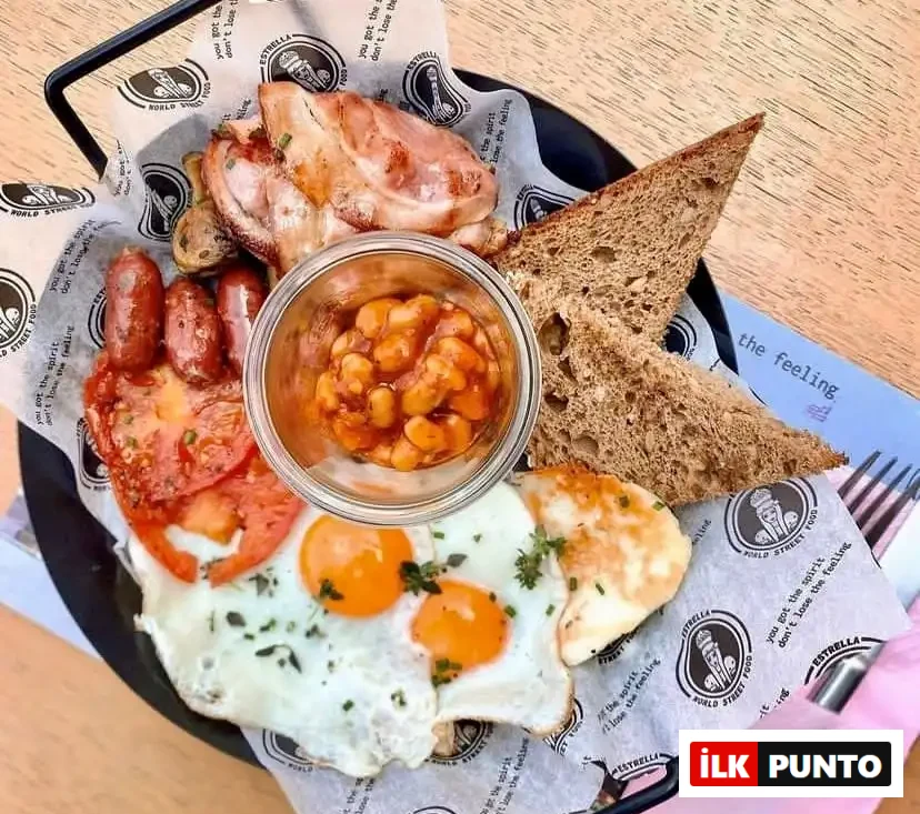 Selanik'in en iyi kahvaltı mekanlarından Estrella'da sunulan, tabağında kızarmış yumurtalar, sucuklar, domates, bacon, fasulye, kızarmış ekmek ve ızgara peynir içeren doyurucu ve bol porsiyonlu bir kahvaltı tabağı.