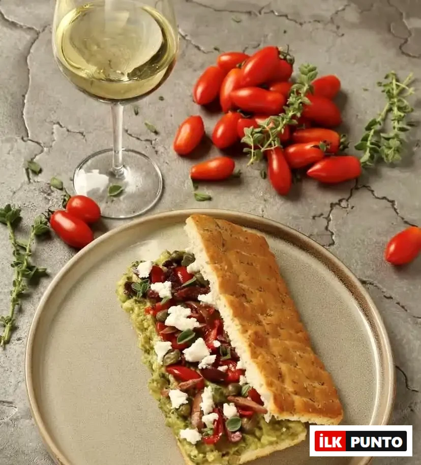 Gri bir zeminin üzerinde duran tabakta, yeşil avokado veya benzeri bir ezme, ufalanmış beyaz peynir, kırmızı biber ve kapari ile zenginleştirilmiş, üzerine taze kekik serpilmiş modern bir ekmek arası sunum yer alıyor; yanında bir kadeh beyaz şarap ve bolca taze çeri domates bulunan bu kare, Selanik'in çeşitli ve çağdaş gastronomik lezzet duraklarından birine ait, iştah açıcı bir öğünü gözler önüne seriyor.