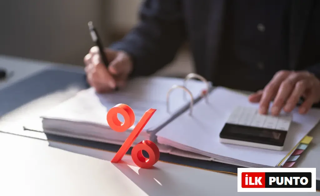 Finansal analiz ve reel faiz hesaplamaları bağlamında, bir masa üzerinde açık bir defterdeki belgelere not alan ve hesap makinesi kullanan kişinin elleri ile ön planda duran belirgin turuncu yüzde işareti.
