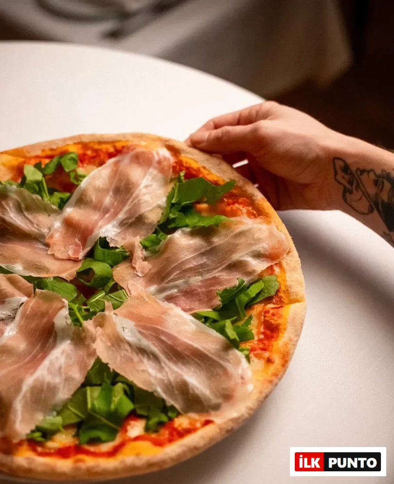 Beyaz bir masa üzerinde, kenarından bir elin tuttuğu, domates sosu, bol yeşil roka ve ince dilimlenmiş prosciutto ile zenginleştirilmiş, nar gibi kızarmış kenarlı yuvarlak bir pizzanın yakın çekimi, Pera gibi bir pizzacının sunduğu özel bir lezzeti yansıtıyor.
