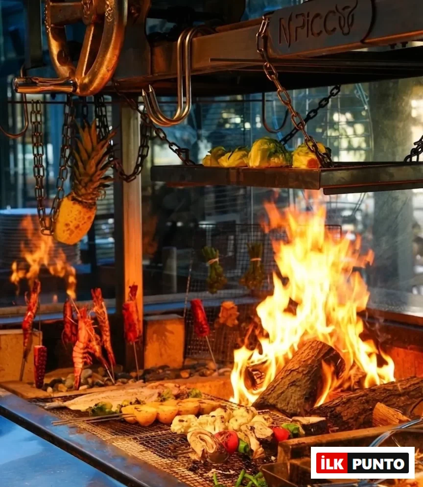 Bir restoranda odun ateşiyle yanan büyük bir mangalda, üst raflarda yeşil sebzeler, zincirlerle asılı yarım ananas, şişlerde karidesler ve kırmızı biberler, ızgara yüzeyinde ise bütün bir balık, patates, brokoli ve çeşitli diğer sebzeler lezzetli bir şekilde pişiriliyor.