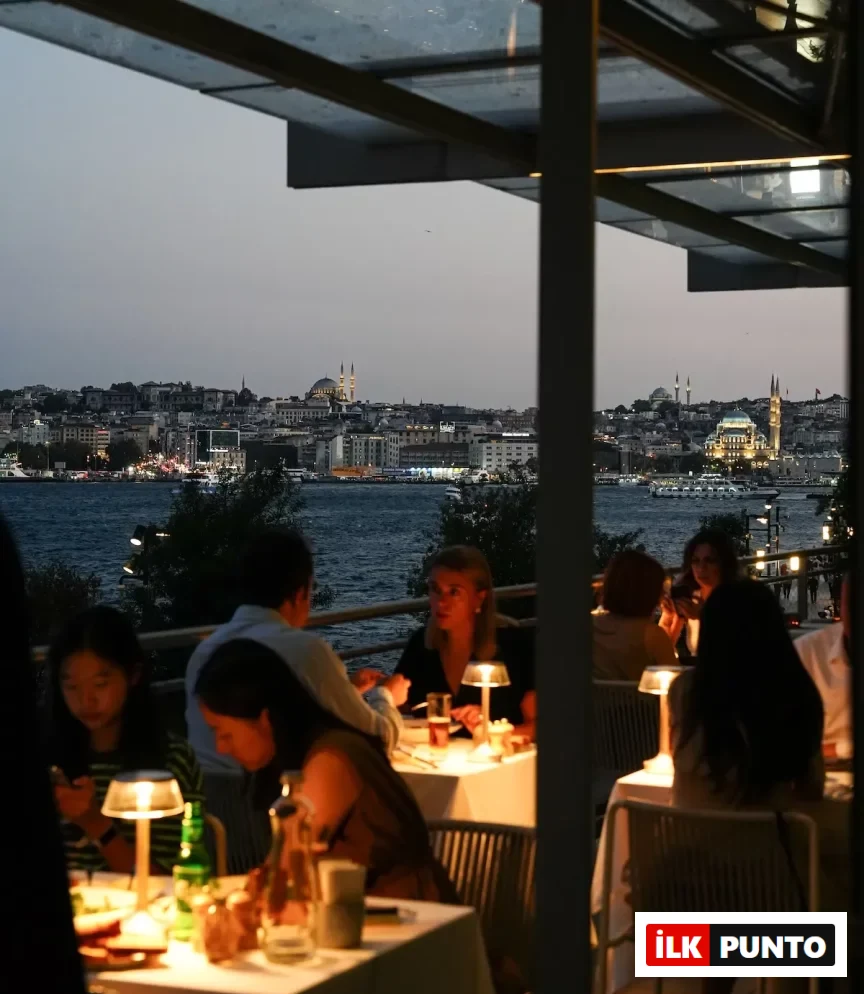 Boğaz manzaralı modern bir restoranda, akşamüstü alacakaranlıkta parıldayan İstanbul silüeti ve tarihi camilerin ışıkları önünde, masalarındaki sıcak lambaların aydınlattığı zarif ortamda yemek yiyen veya sohbet eden misafirler.