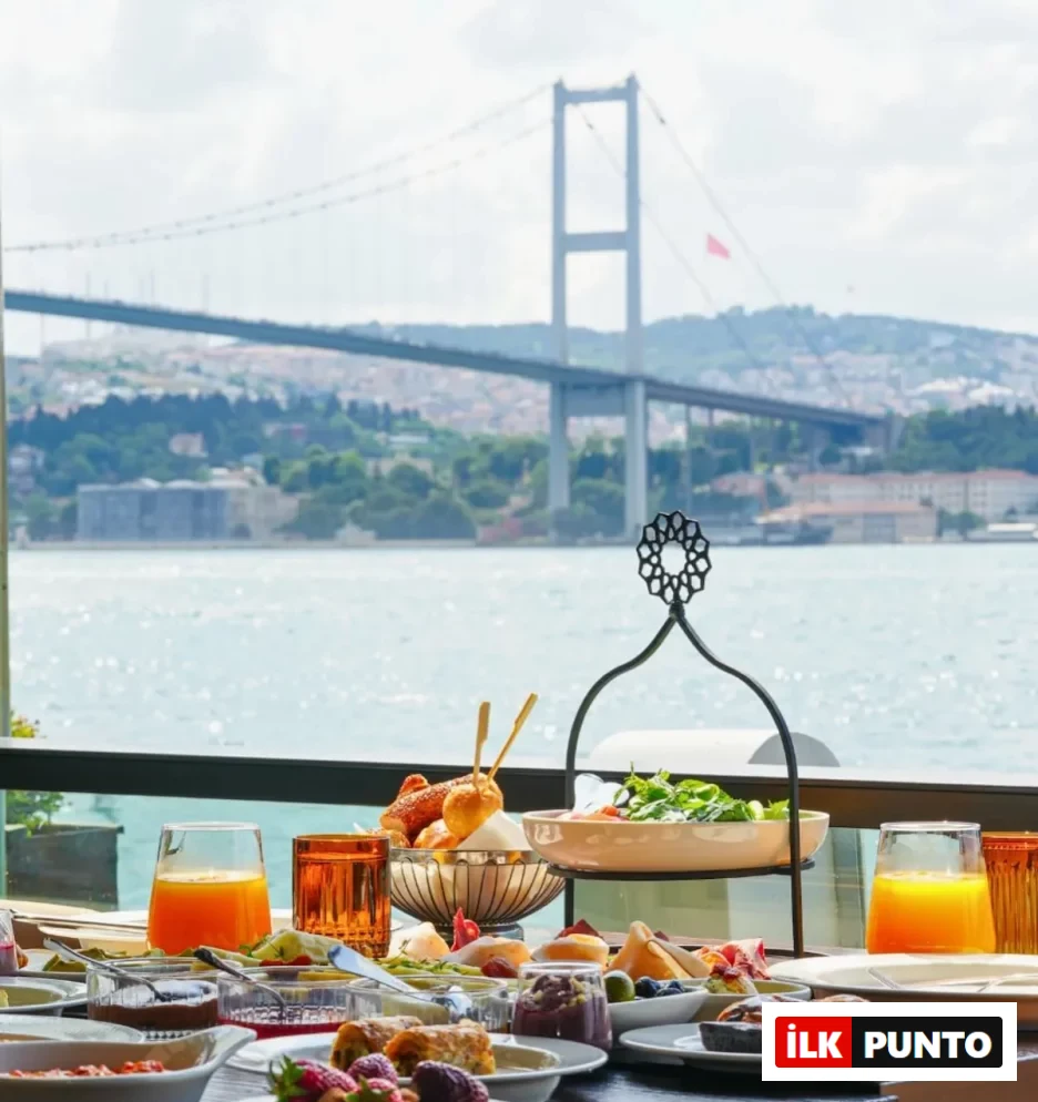 Özenle hazırlanmış, çilekler, böğürtlenler, taze hamur işleri, reçeller, salata ve iki bardak taze sıkılmış portakal suyunun bulunduğu zengin bir kahvaltı sofrası, arkasında net bir şekilde İstanbul Boğazı'nın ve etkileyici Boğaziçi Köprüsü'nün göz alıcı manzarasıyla birlikte konumlanmış.