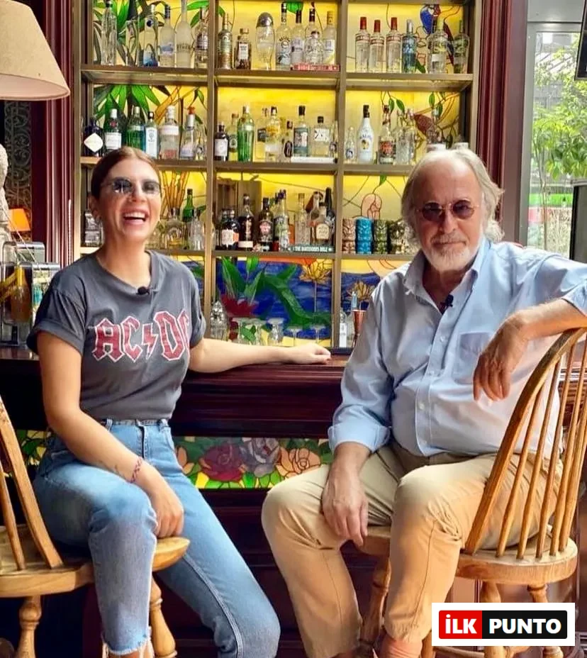 Bir bar taburesinde oturan, AC/DC tişörtlü güler yüzlü bir kadın ve açık mavi gömlekli, sakallı bir adam, arkalarında renkli vitray detayları ve çeşitli şişelerle dolu bir raf ünitesi bulunan canlı bir barda keyifli bir anı paylaşıyorlar.