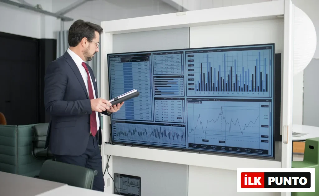 Takım elbiseli bir adam, finansal verilerin ve grafiklerin gösterildiği büyük bir ekrana analiz amaçlı bakıyor.