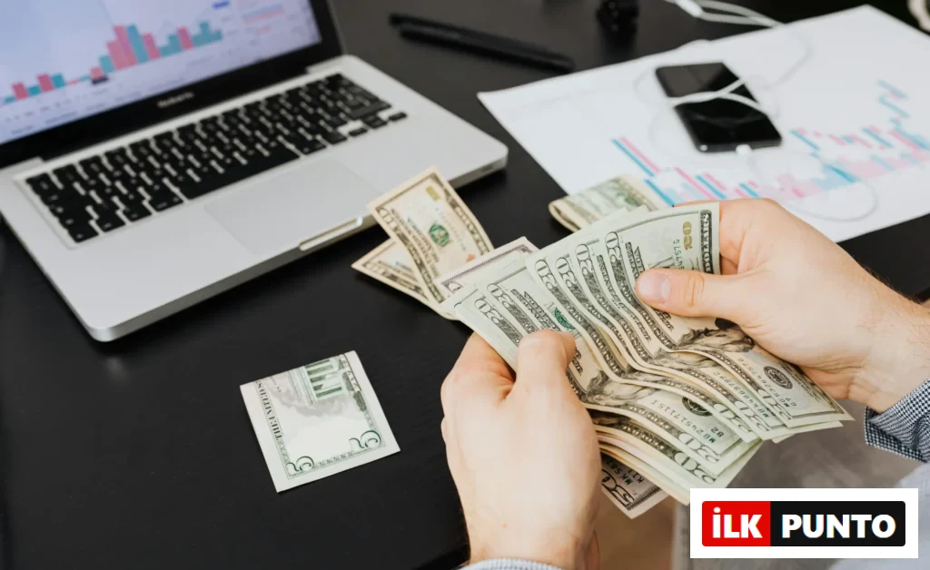 Bir kişi, masa üstünde laptop ve grafikler varken elindeki Amerikan dolarlarını sayıyor.
