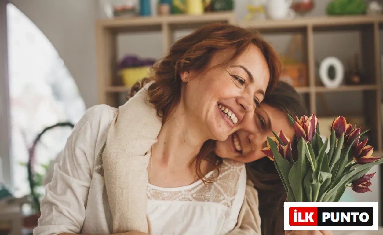 Gülümseyen, mutlu bir anne ve ona sarılarak başını omzuna yaslamış genç kızı, kızının elinde tuttuğu canlı kırmızı ve sarı tonlarında lale buketiyle neşeli ve sıcak bir anı paylaşıyorlar.